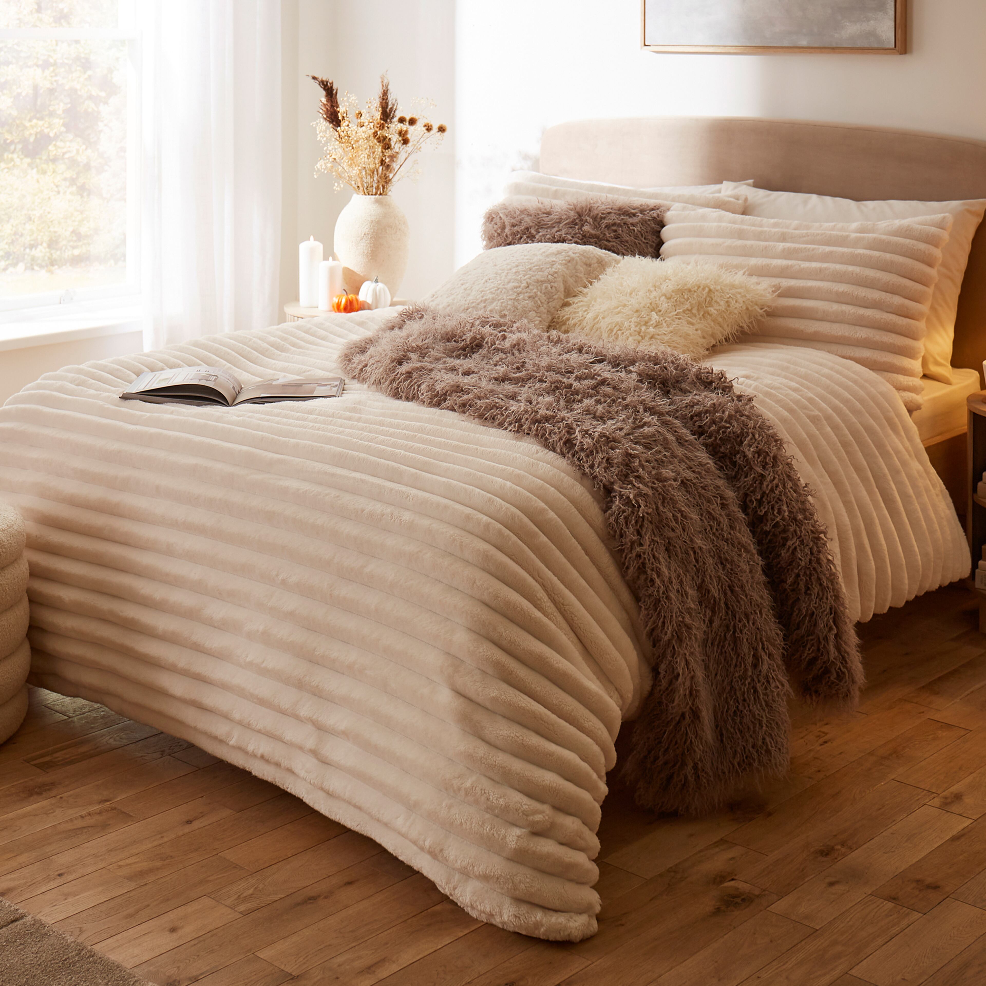 Cosy bedroom with a beige upholstered bed, cream striped bedding, fluffy throw blanket, and decorative pillows. 